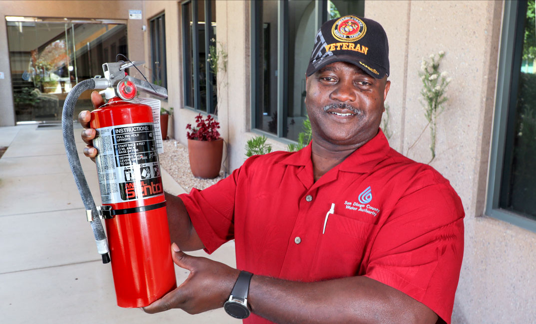 An employee standing in front of the Kearny Mesa building wearing a Marine Corps veteran cap, a Water Authority logo shirt and holding a fire extinguisher.