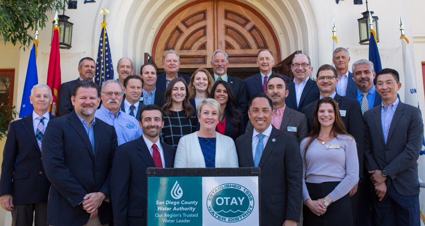 A group of regional elected officials and water industry leaders and their staff stand behind a podium with the Water Authority and Otay Water District logos.